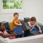 Children playing on a foam pit in a room with a window showing greenery outside.