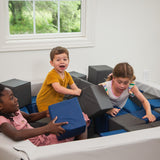 Children playing on a foam pit in a room with a window showing greenery outside.
