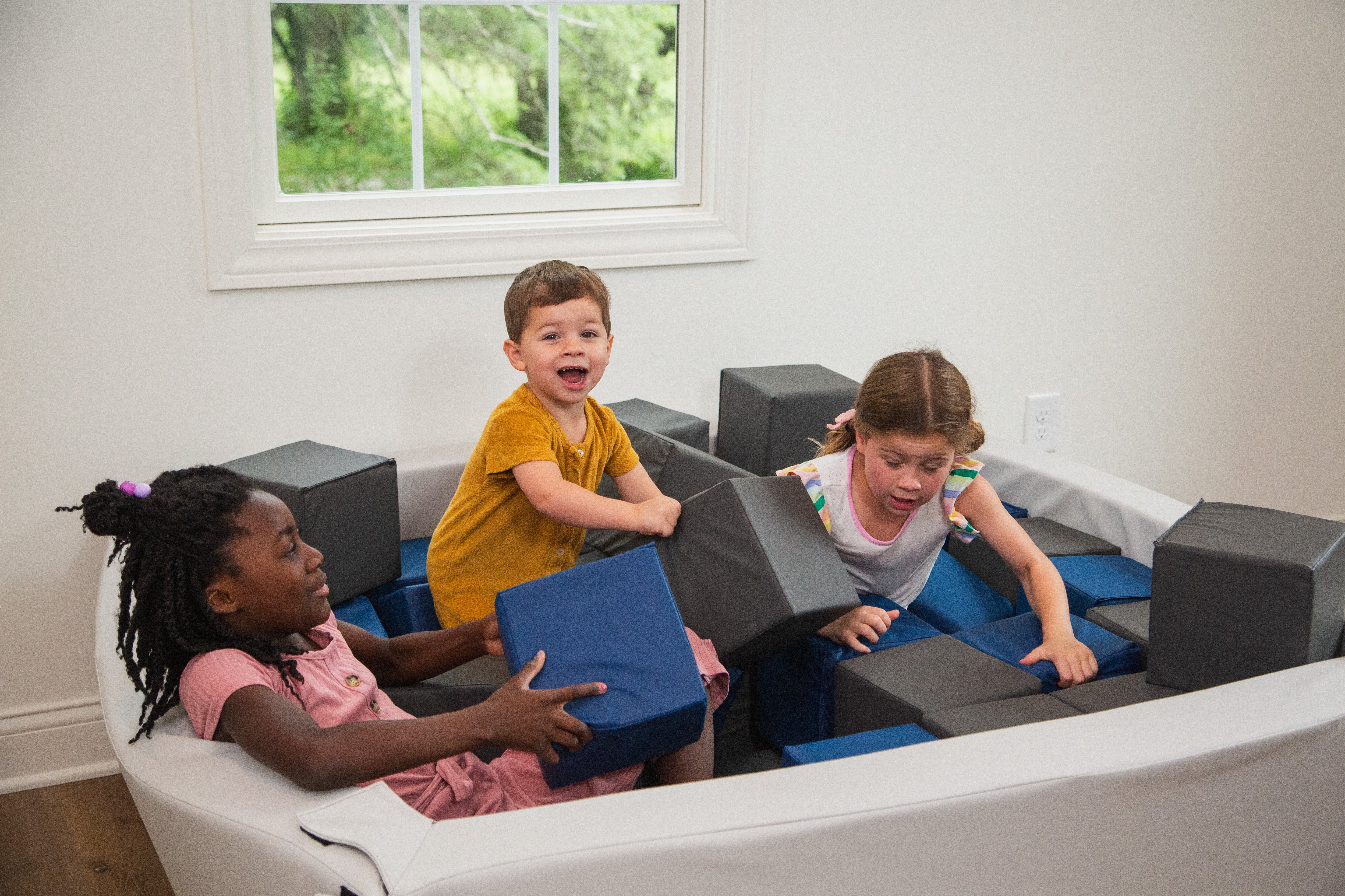 Children playing on a foam pit in a room with a window showing greenery outside.