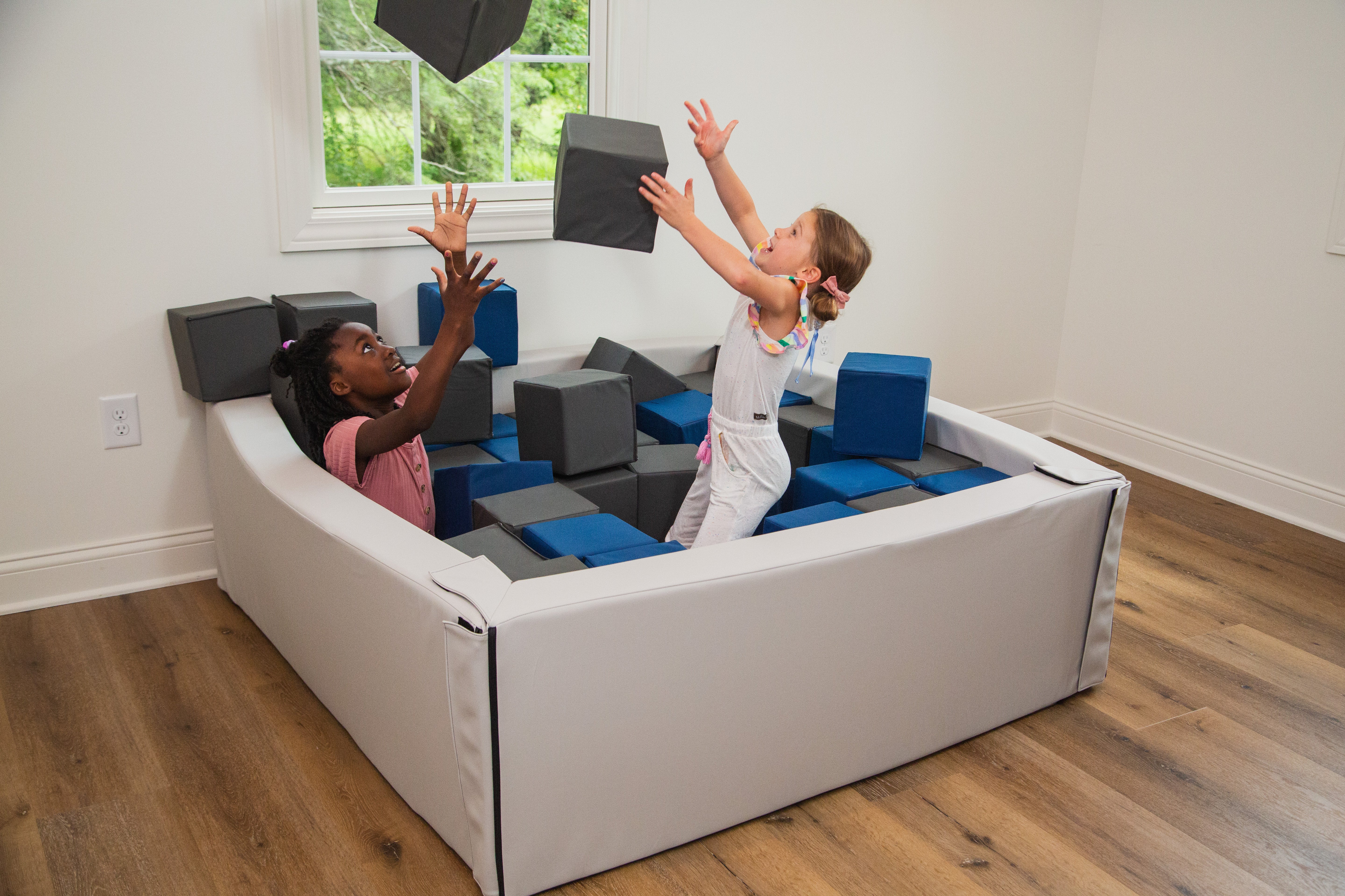 Two children playing with soft play blocks in a room with wooden flooring and large windows.