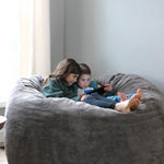 Two children sitting on a large Haven bean bag chair, looking at a device.