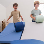 Two kids rolling foamnasium cylinders down mega wedges in living room 