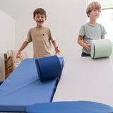 Two kids rolling foamnasium cylinders down mega wedges in living room 