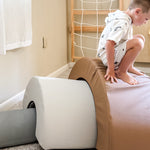 A child in a white hoodie and shorts crouches on a large tan and pink foam balance block in a playroom, with a gray foam block, wooden climbing wall, and carpeted floor visible.