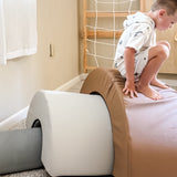 A child in a white hoodie and shorts crouches on a large tan and pink foam balance block in a playroom, with a gray foam block, wooden climbing wall, and carpeted floor visible.