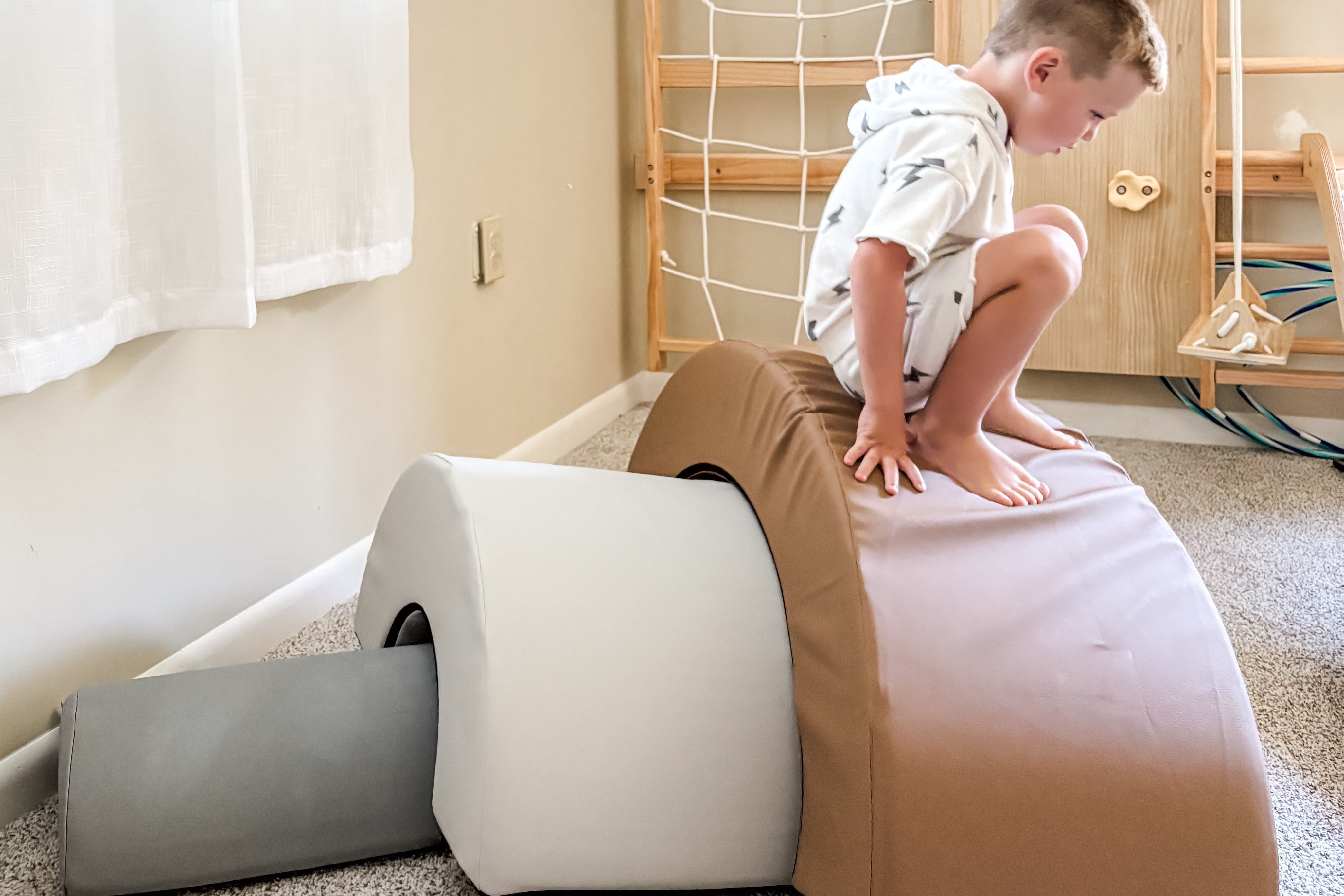 A child in a white hoodie and shorts crouches on a large tan and pink foam balance block in a playroom, with a gray foam block, wooden climbing wall, and carpeted floor visible.