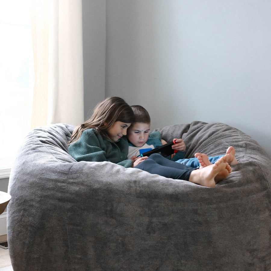 Two children sitting on a large Haven bean bag chair, looking at a device.